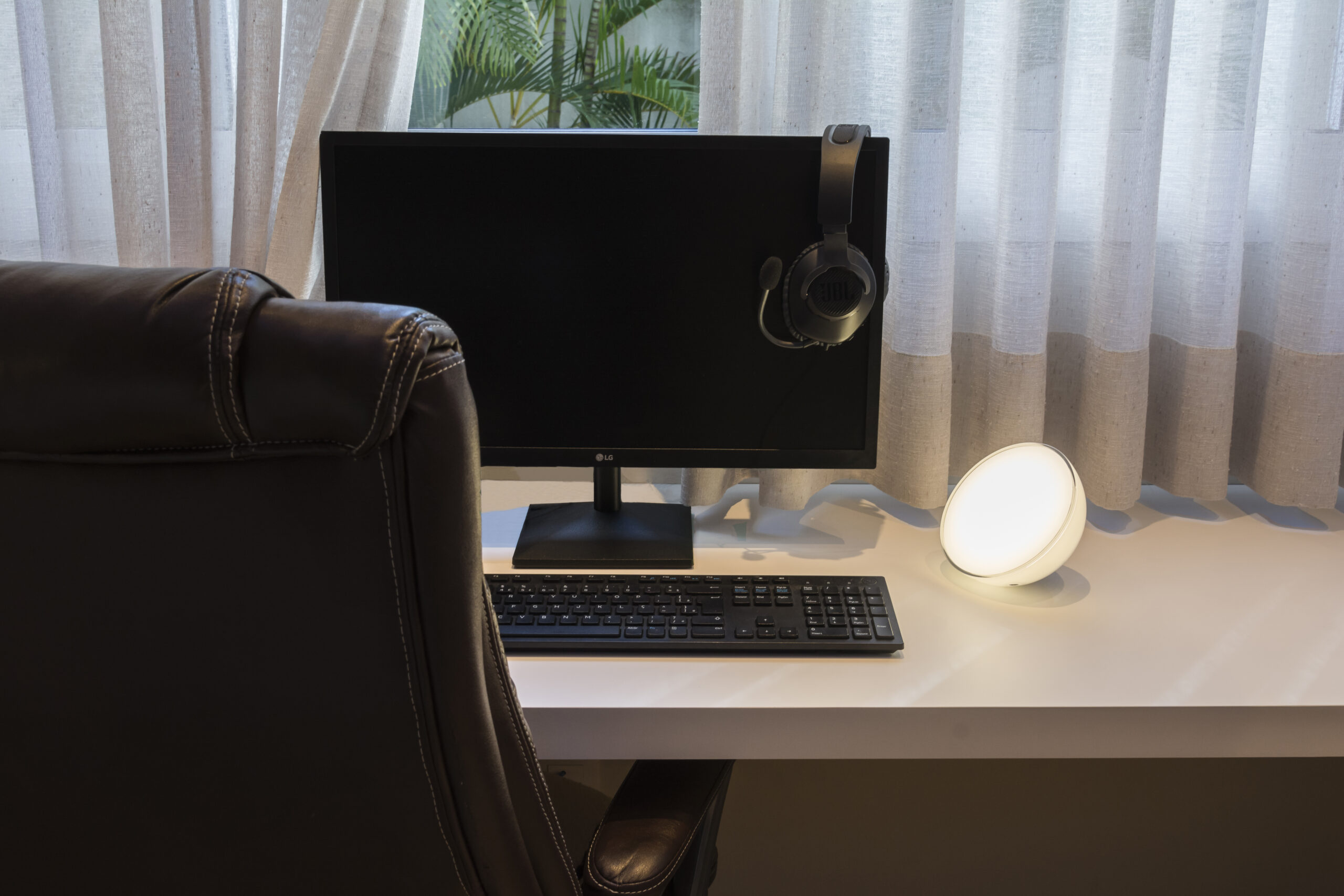 Escrivaninha de madeira branca com monitor, teclado e headset, cadeira de escritório vista de costas, luminária de mesa apontada para o monitor e, ao fundo, uma janela aberta com cortina branca entreaberta.