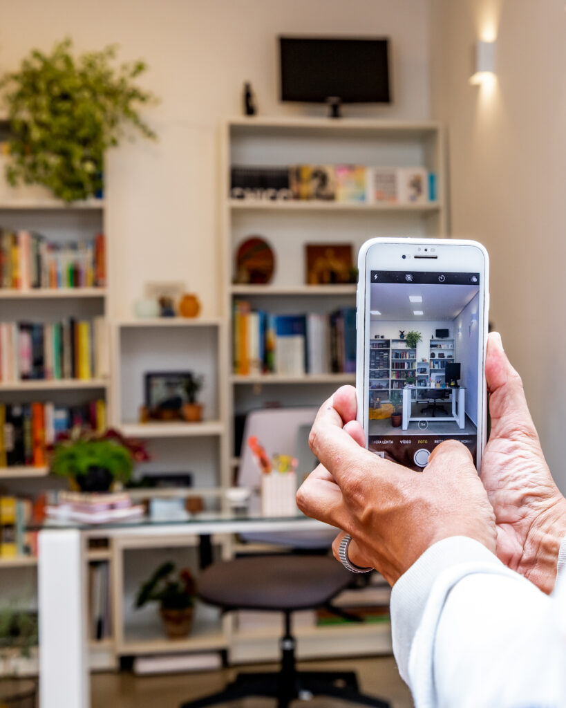 Mãos de uma pessoa segurando um celular que está tirando uma foto de um home office com diversas prateleiras e uma mesa brancas. É possível ver plantas e objetos decorativos e muitos livros espalhados pelo ambiente. 