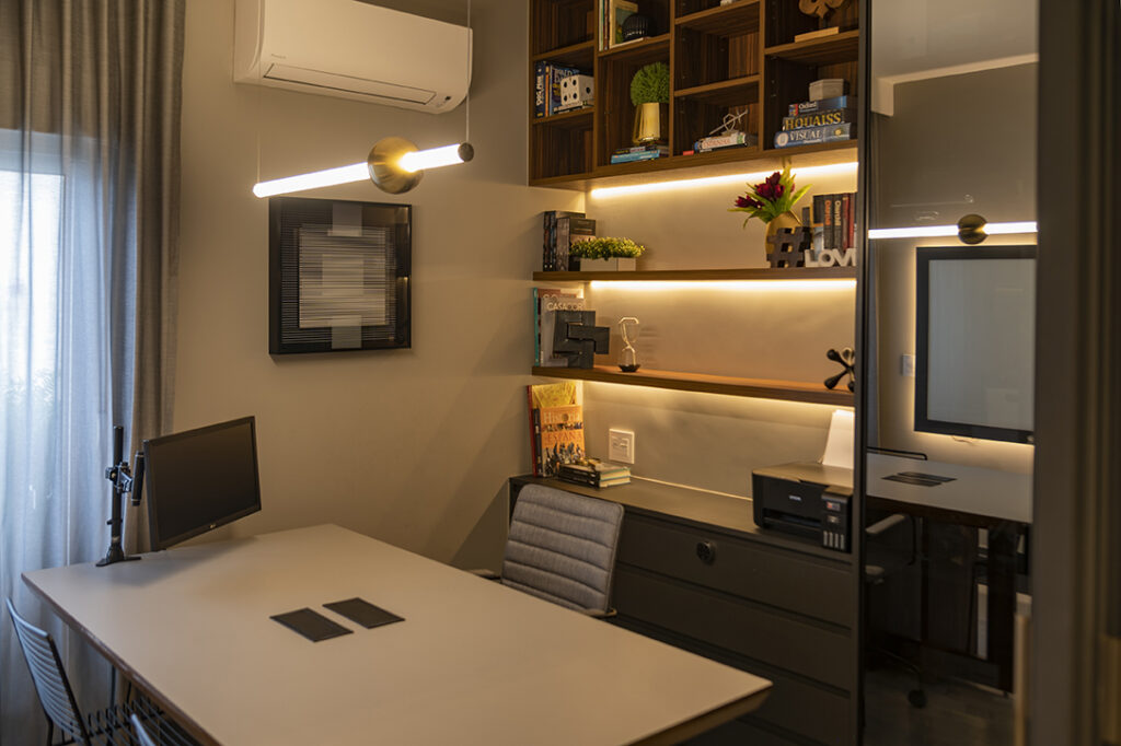 Home office mobiliado em madeira e iluminado com luminária pendente de led e fitas de led nas prateleiras que ficam atrás da mesa. Vemos plantas decorativas e livros nas prateleiras. Vemos um quadro e um aparelho de ar condicionado na parede. 