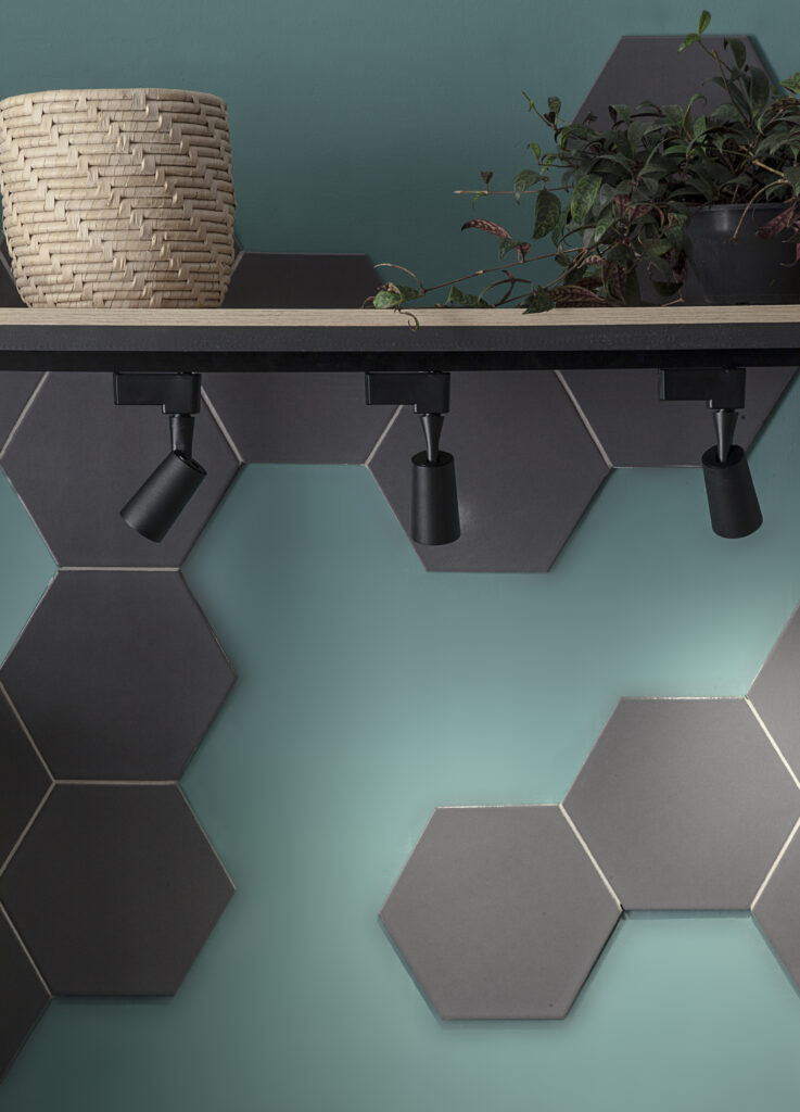 Área de serviço moderna com algumas plantas decorativas e cestos com materiais de limpeza e uma calha com 3 spots pretos. Parede verde com revestimento em mosaico hexagonal cinza.