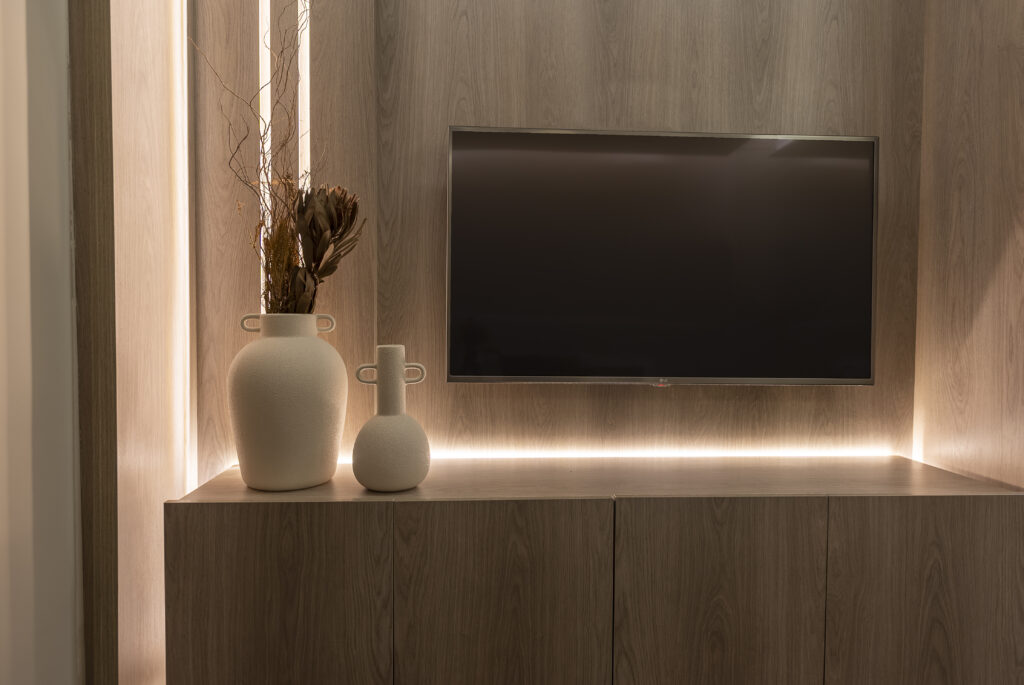 Na sala minimalista, a TV de tela plana está fixada na parede de madeira, logo acima de um gabinete. Ao lado, um vaso com plantas secas adiciona textura, enquanto um pequeno objeto esférico chama a atenção. A iluminação parece ser toda feita com fitas de led