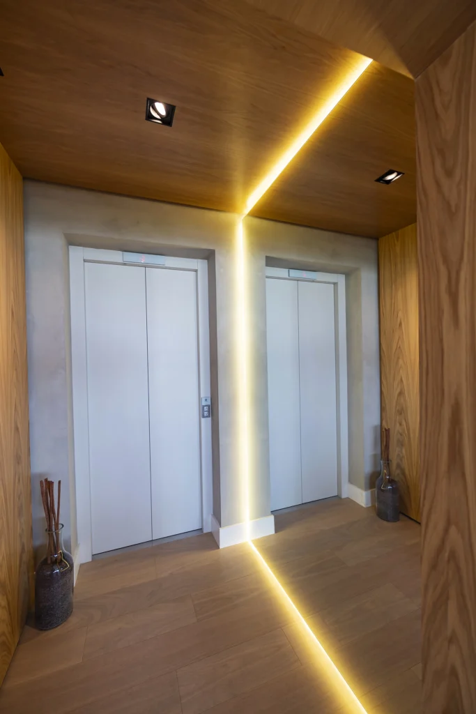 Interior de hall de entrada moderno com uma porta de elevador branca fechada, piso e paredes de madeira e espelho de parede com iluminação embutida de fitas de led criando uma linha amarela ao seu redor.