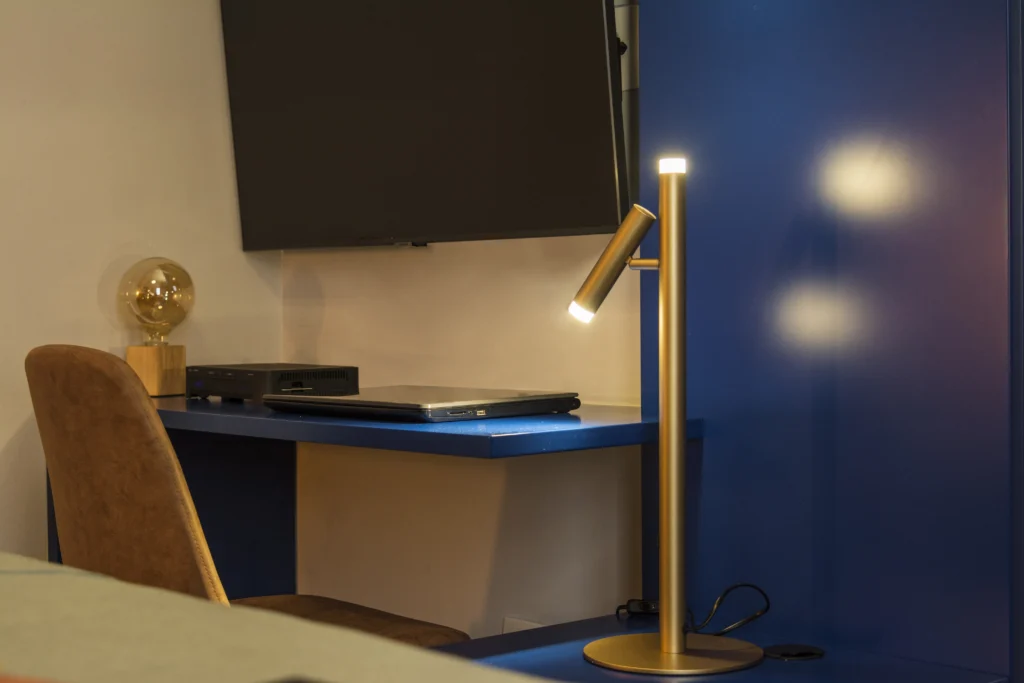 Uma mesa de estudo azul com uma cadeira marrom, sobre a qual estão um laptop fechado, um roteador e uma lâmpada decorativa com bulbo de vidro. À direita, uma luminária para mesa de estudo moderna e dourada, com duas luzes acesas, ilumina o ambiente. Acima da mesa, uma televisão está fixada na parede, adicionando um toque tecnológico ao espaço.