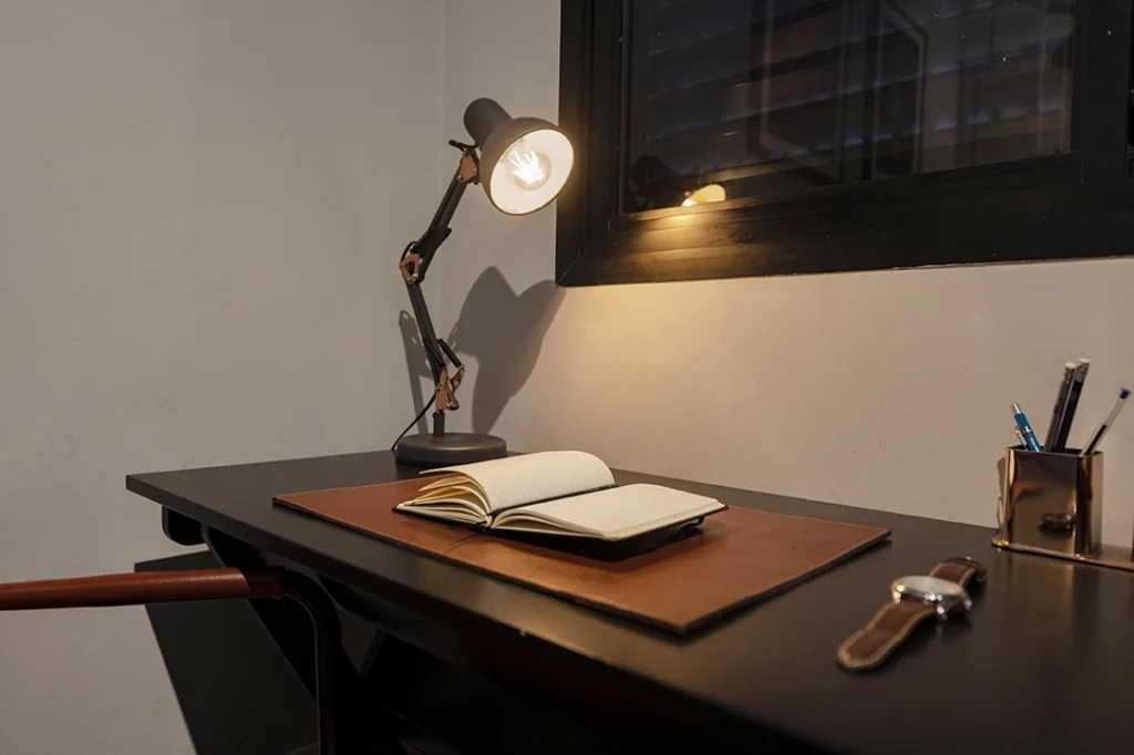 A imagem mostra uma luminária para mesa de estudo articulada acesa sobre uma mesa de estudo escura com uma. Na mesa, há um caderno aberto sobre um suporte de couro marrom. À direita, um porta-lápis metálico contém alguns lápis e canetas, e à frente dele, um relógio de pulso com uma pulseira de couro está colocado sobre a mesa. Ao fundo, uma janela com venezianas pretas permite a entrada de luz. O ambiente parece organizado e ideal para estudo ou trabalho.