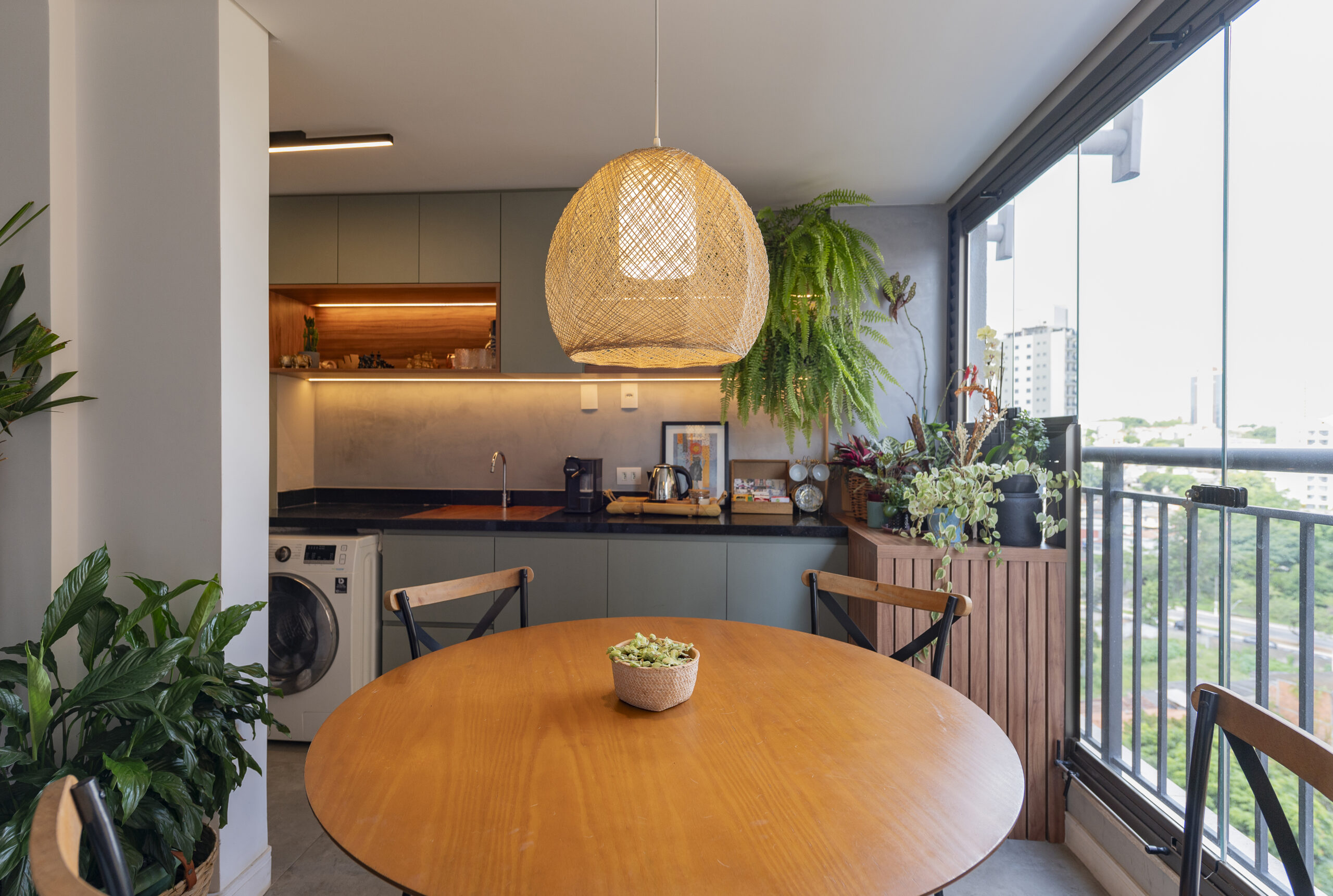 Sala de jantar contemporânea com peças em madeira clara e lustre com abóboda em vime. Ao fundo vemos uma janela que parece dar para uma varanda e plantas decorativas.