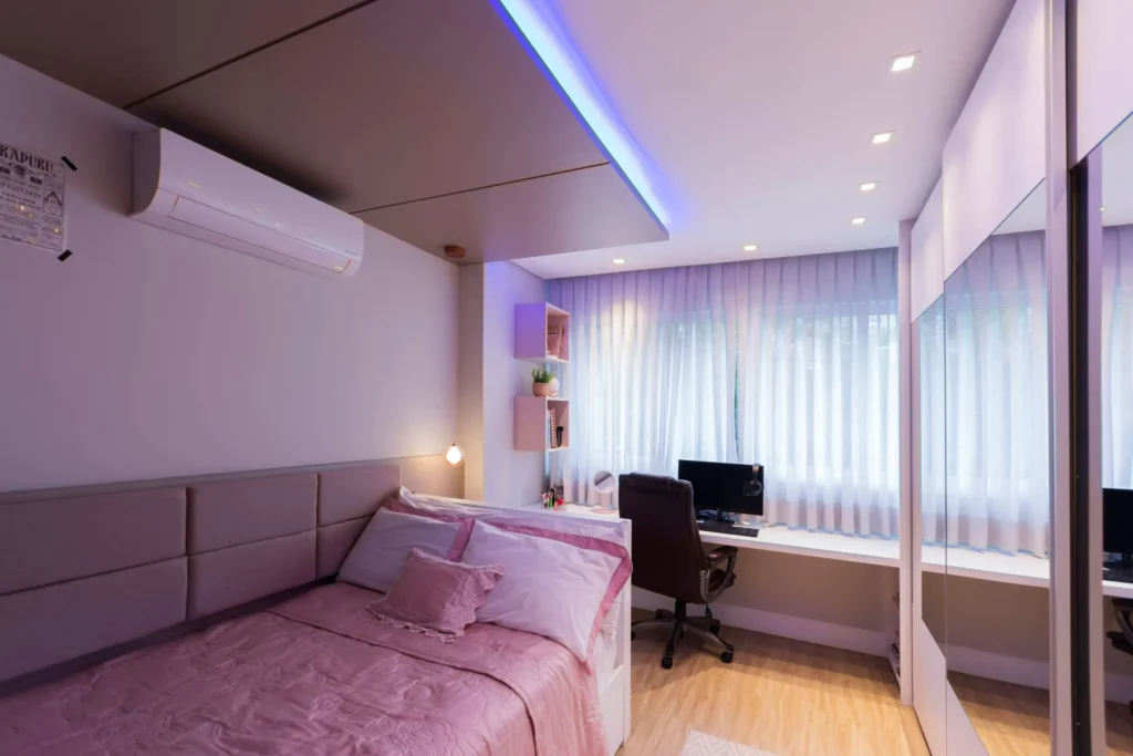 Quarto moderno com cama de de solteiro, roupas de cama rosas, cortinas brancas, iluminação azul indireta feita com fita de led, ar-condicionado e mesa de trabalho com computador.