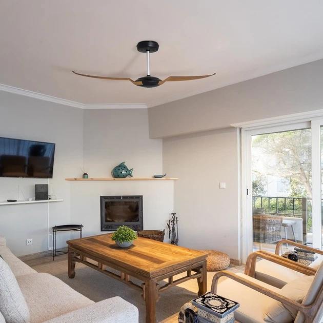 Sala de estar acolhedora com ventilador de teto preto e pás em tom de caramelo, combinando com a mesa de centro de madeira rústica. O ambiente é decorado com sofá bege, poltronas confortáveis, lareira moderna e uma porta de vidro que se abre para uma varanda com vista para a natureza.