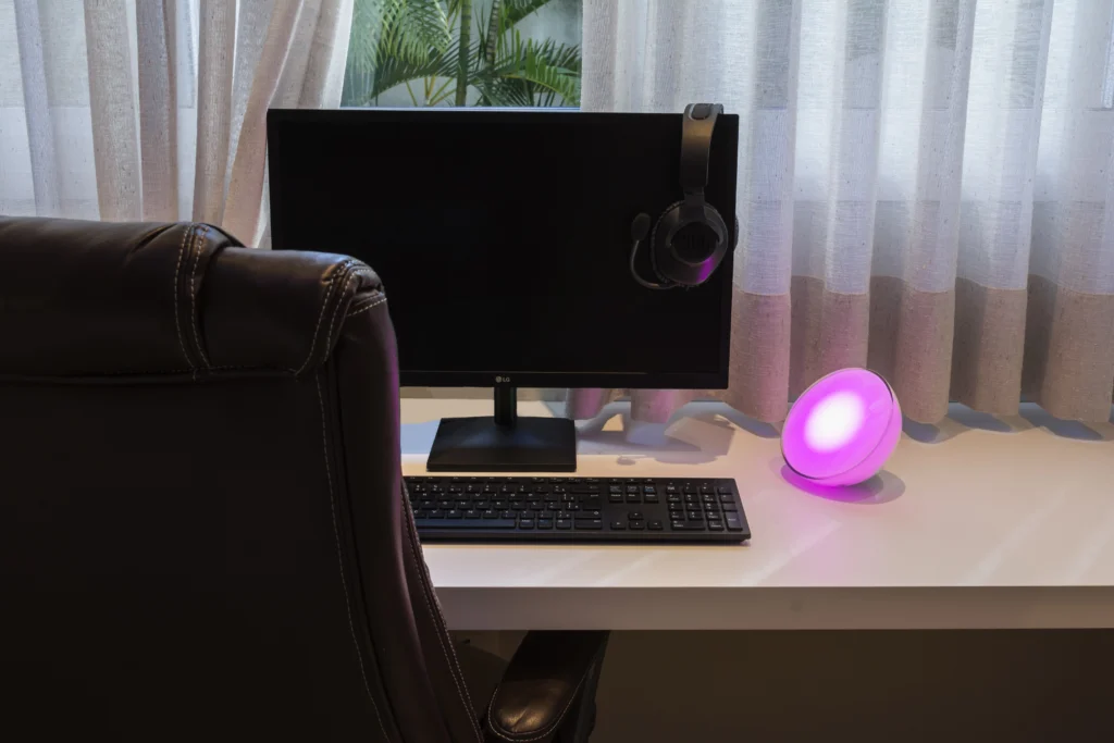 Mesa de setup gamer com monitor, teclado e fone de ouvido, destacada por uma luminária de LED roxa, criando um ambiente moderno e aconchegante para jogos.