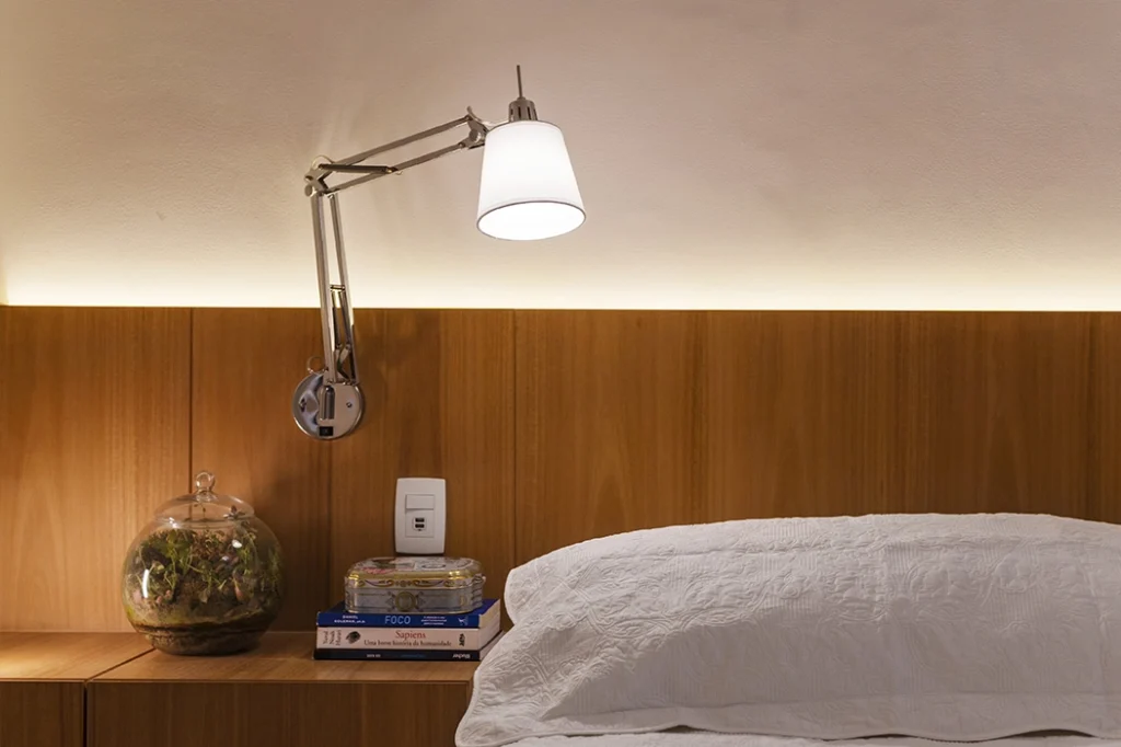 Luminária de parede ajustável ao lado de uma cama com cabeceira de madeira, destacando o sistema de dimerização, ideal para controle de intensidade de luz em ambientes aconchegantes, como um quarto.