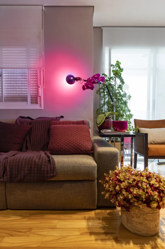 Sala de estar aconchegante com sofá cinza decorado com almofadas e manta em tons de roxo, iluminada por uma luminária de parede emitindo luz rosa, complementada por plantas decorativas e uma mesa lateral moderna.