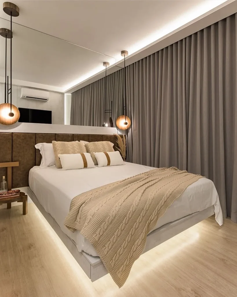 A imagem mostra um quarto de casal moderno e elegante. O ambiente é iluminado por luzes embutidas no teto, criando uma atmosfera aconchegante. A cama, central na imagem, é coberta por roupas de cama brancas e decorada com almofadas e uma manta em tons neutros, como bege e creme. A cabeceira da cama é alta e estofada, com um painel espelhado acima, refletindo parte do quarto e ampliando visualmente o espaço. Pendentes modernos com globos de vidro e estrutura em metal escuro estão suspensos ao lado da cama, proporcionando iluminação suave. Um ar-condicionado split está instalado na parede ao fundo, garantindo conforto térmico. Ao lado da cama, uma mesa de cabeceira em madeira natural complementa a decoração. O chão é de madeira clara, e uma iluminação indireta sob a cama destaca o piso, acrescentando um toque de sofisticação ao ambiente. Cortinas longas em um tom de cinza suave cobrem as janelas, completando o design do quarto.