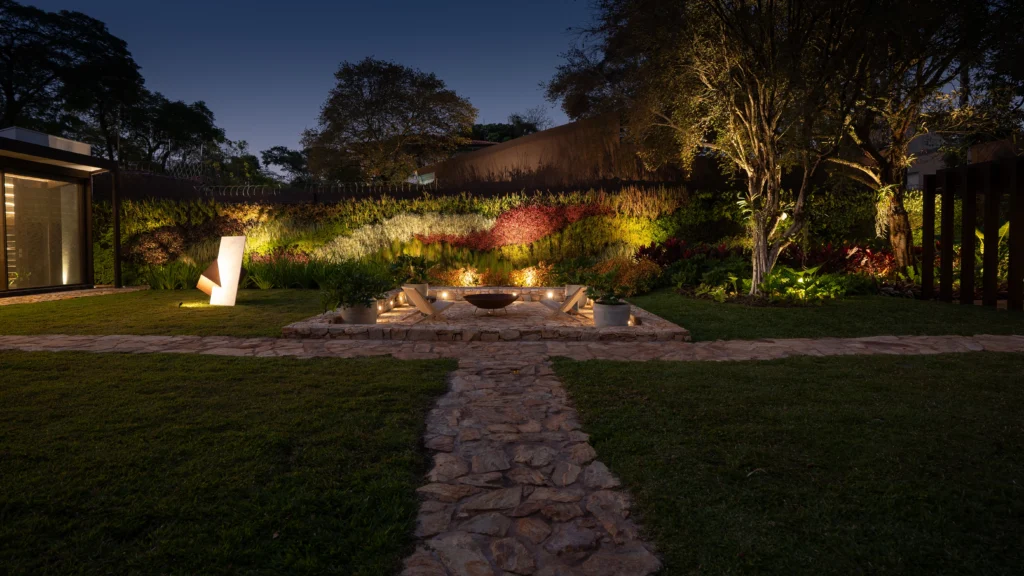 <img src="jardim-iluminacao-noturna.webp" alt="Vista noturna de um jardim com iluminação suave, caminho de pedras e áreas verdes exuberantes, ideal para projetos de paisagismo e decoração exterior.">