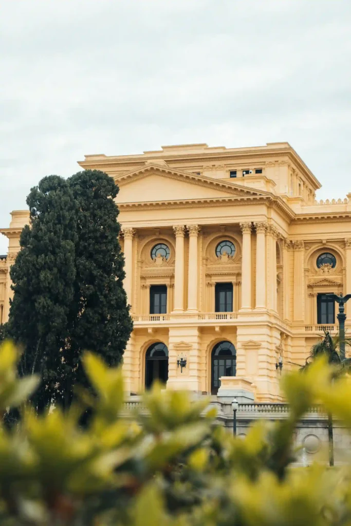 <img src="predio-historico-arquitetura-amarela.webp" alt="Vista do prédio histórico em tom amarelo, com detalhes arquitetônicos, janelas grandes e elementos decorativos, rodeado por árvores verdes.">