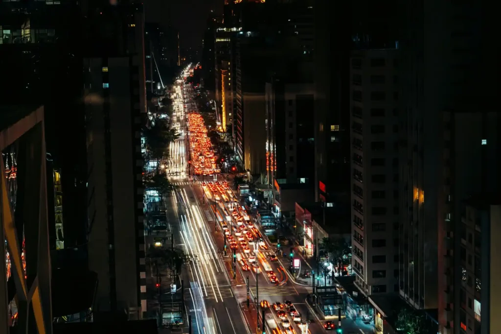 <img src="vista-noturna-trafego-intenso.webp" alt="Vista noturna da cidade com tráfego intenso, onde as luzes dos carros criam um efeito vibrante na rua. Ideal para ilustrar a vida urbana agitada.">