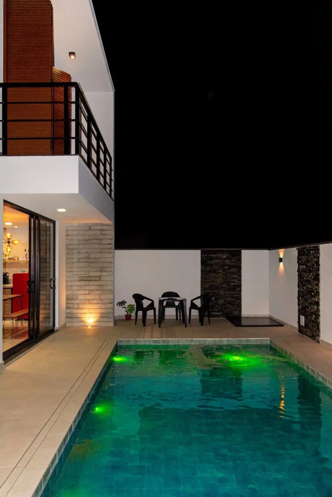 <img src="piscina-iluminada-com-area-de-churrasco-e-area-de-estar-ao-ar-livre-jpg.webp" alt="Imagem de uma piscina iluminada com luzes verdes ao lado de uma área de churrasco ao ar livre, incluindo uma área de estar com cadeiras pretas, parede de pedra e casa ao fundo, durante a noite.">