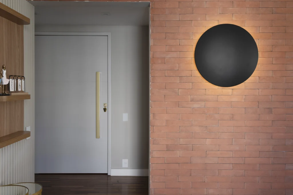 <img src="interior-sala-moderno-parede-tijolos-espelho-preto.webp" alt="Interior de uma sala moderna com parede de tijolos aparentes, destaque para um espelho circular preto iluminado e uma porta branca com puxador longo.">