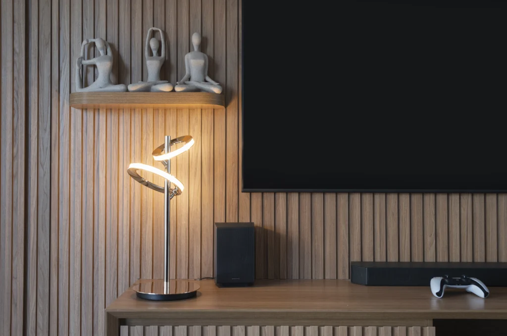 <img src="decoracao-modena-sala-madeira-luminaria-design.webp" alt="Decoração moderna de sala com painel de madeira, luminária de design exclusivo, TV e dispositivos eletrônicos em um ambiente aconchegante.">