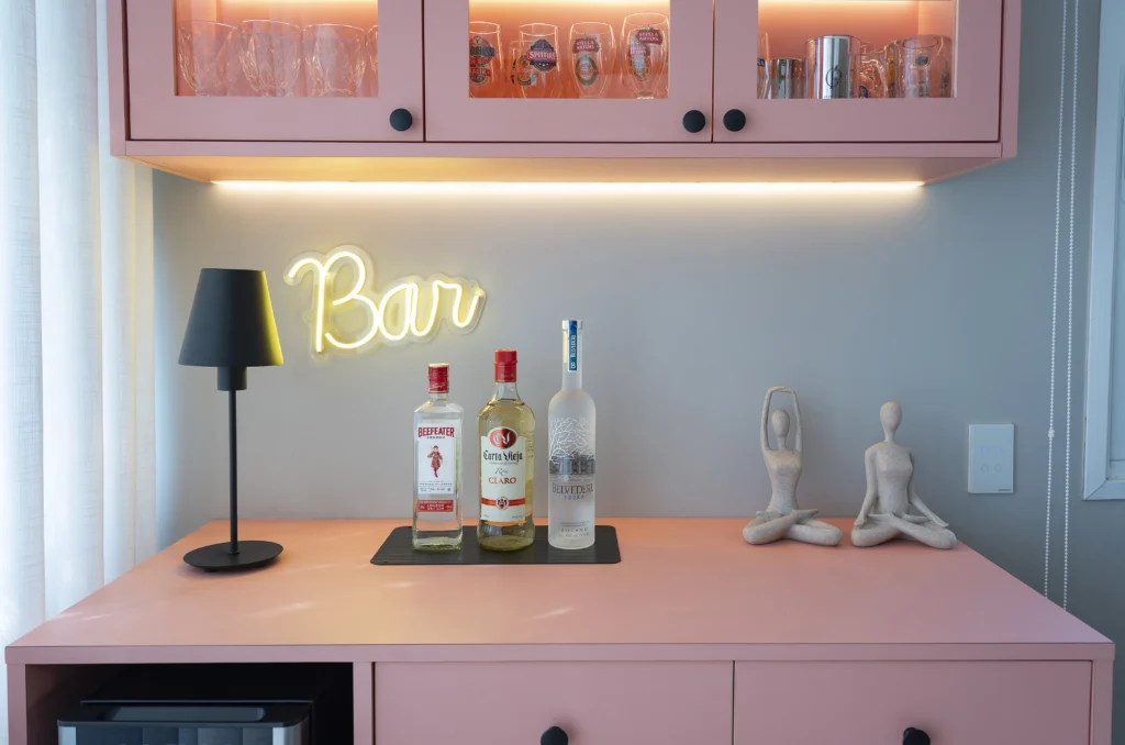 <img src="decoracao-moderna-de-bar-com-luz-neon.webp" alt="Decoração moderna de bar com luz de neon 'Bar', garrafas de bebidas, luminária preta, objetos decorativos e armário rosa claro, criando um ambiente acolhedor e estiloso.">