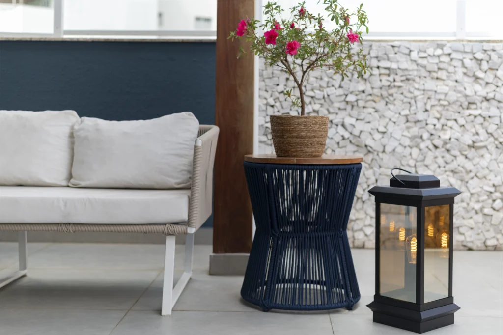 <img src="decoracao-varanda-modernista-com-plantas-e-lanterna-jpg.webp" alt="Ambiente de varanda com sofá branco, mesa de fibra sintética azul, vaso de planta com flores pink e lanterna decorativa, ideal para decoração de varanda moderna.">