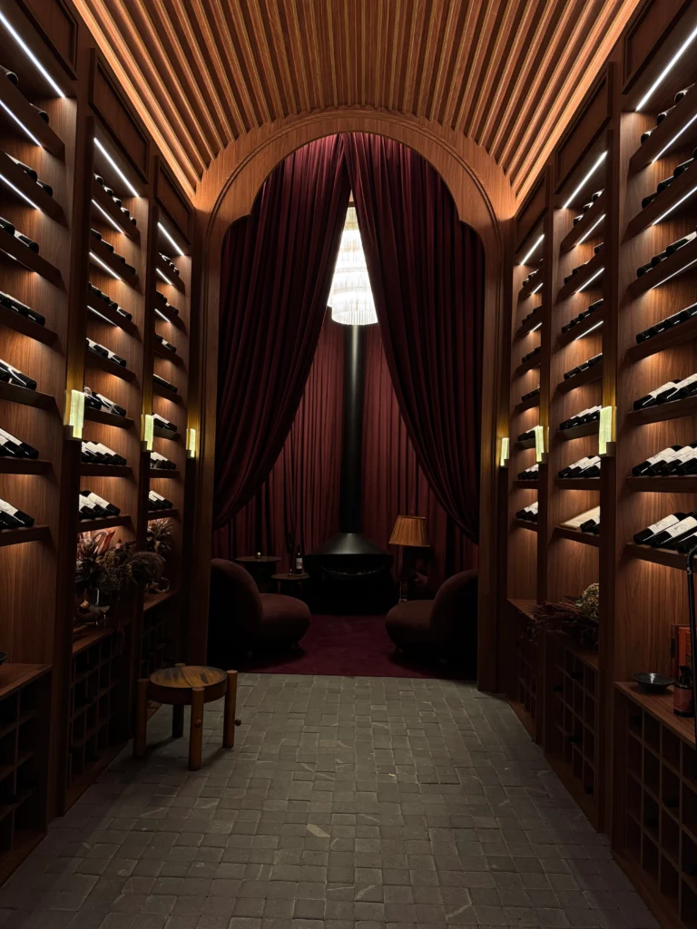 <img src="adega-de-vinho-elegante-com-prateleiras-e-cortinas.webp" alt="Imagem de uma elegante adega de vinho com paredes de madeira, prateleiras com garrafas de vinho e um espaço aconchegante com poltronas, cortinas marcantes e iluminação suave.">