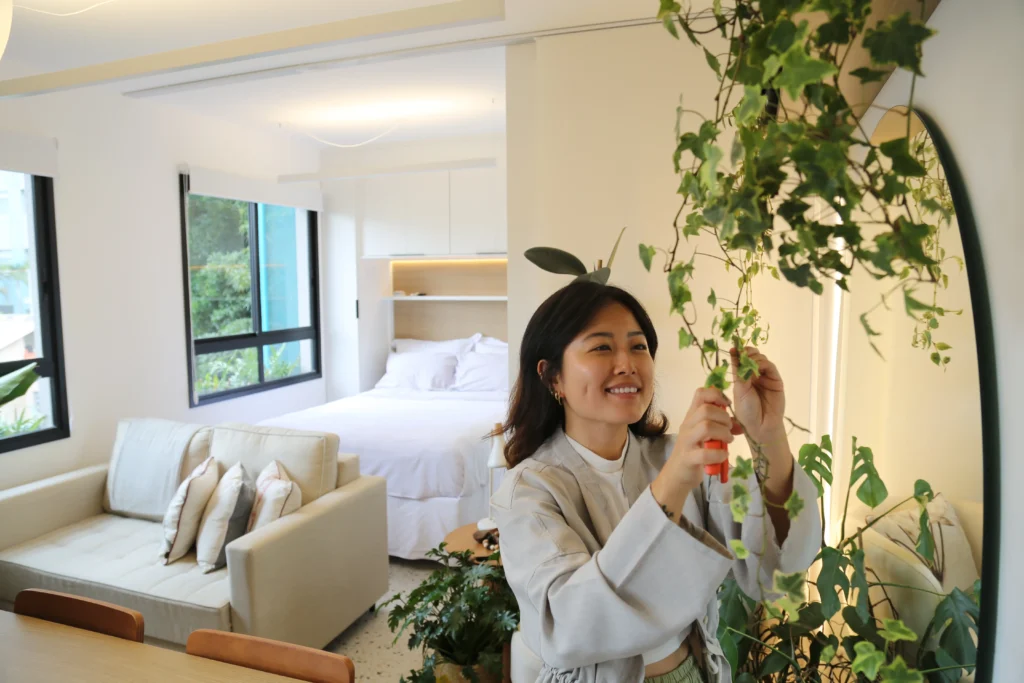 <img src="mulher-sorridente-cuidando-plantas-apartamento-modern.webp" alt="Mulher sorridente cuidando de plantas em um ambiente moderno de apartamento com sala de estar e quarto ao fundo, com foco em sustentabilidade e bem-estar.">