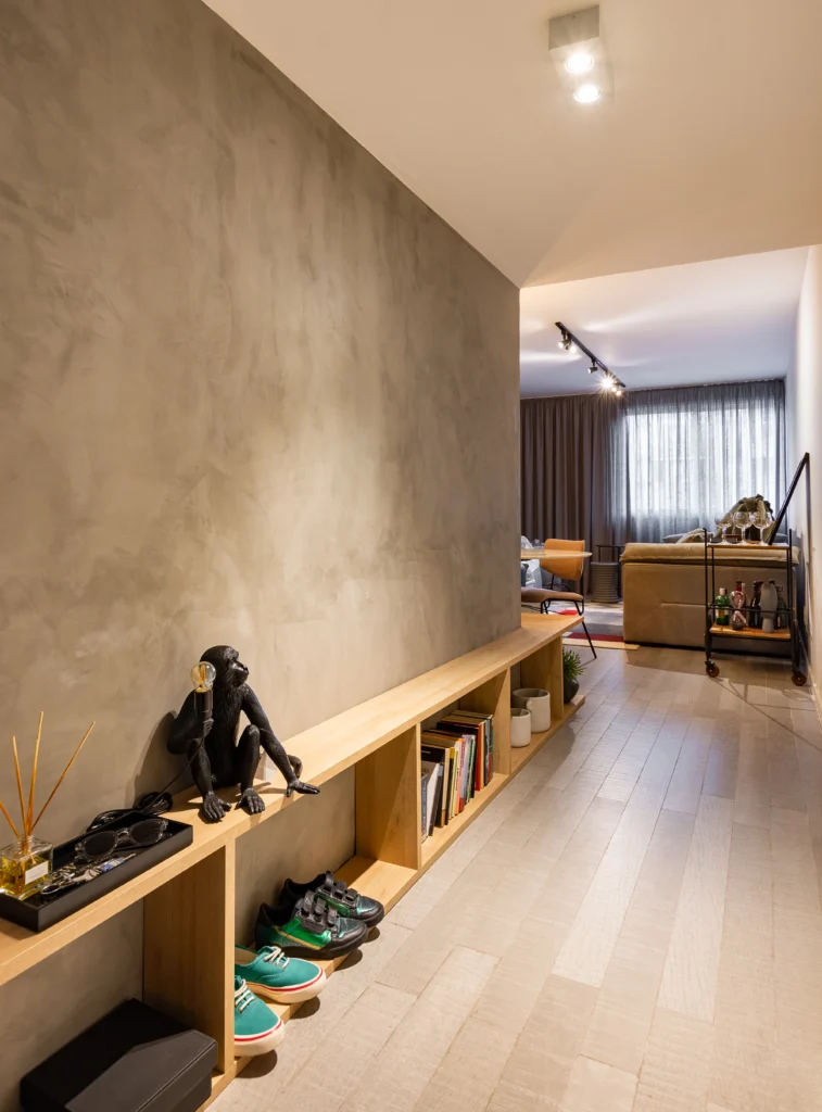 <img src="decoracao-interior-sala-prateleira-objetos.webp" alt="Imagem de um corredor de apartamento com piso de madeira, uma prateleira com livros e objetos decorativos, incluindo uma escultura de macaco com fone de ouvido, na área de estar ao fundo com sofá e cortinas opacas.">
