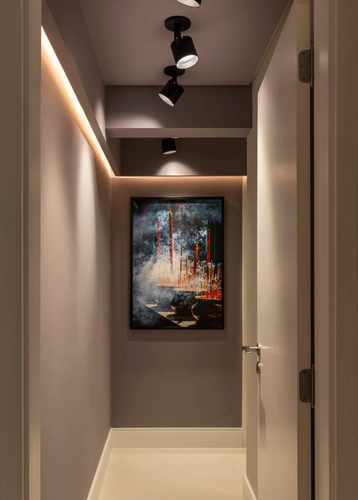 <img src="corredor-com-arte-moderno-iluminacao-embutida.webp" alt="Imagem de um corredor com iluminação embutida e quadro de arte moderna na parede ao fundo, criando um ambiente elegante e contemporâneo.">