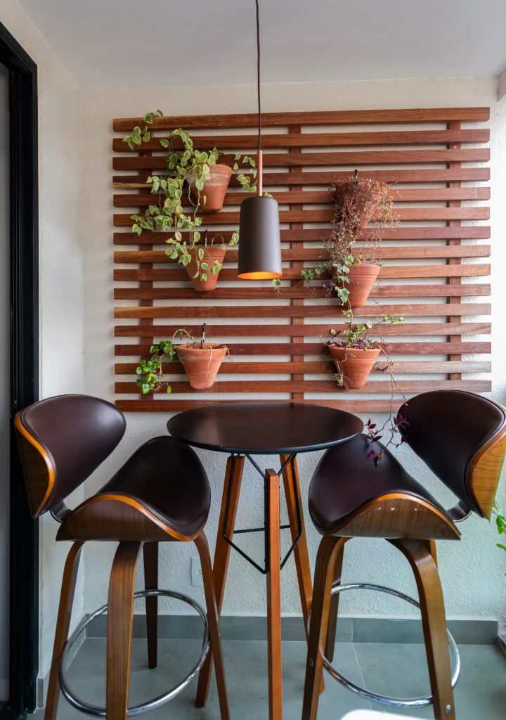<img src="varanda-com-mesa-e-cadeiras-modernas-plantas-e-luz.webp" alt="Decoração de varanda com mesa redonda e cadeiras modernas, parede de madeira com vasos de plantas, iluminação pendente criando ambiente aconchegante.">