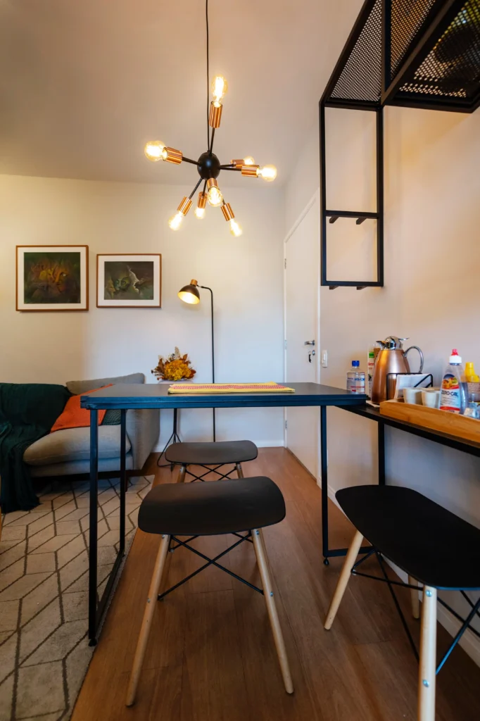 <img src="sala-de-jantar-moderno-com-ustensilios-de-cozinha-e-luminaria.webp" alt="Imagem de uma sala de jantar moderna com mesa preta, banquetas pretas, luminária de teto com lâmpadas, parede decorada com quadros e uma área com prateleiras.">