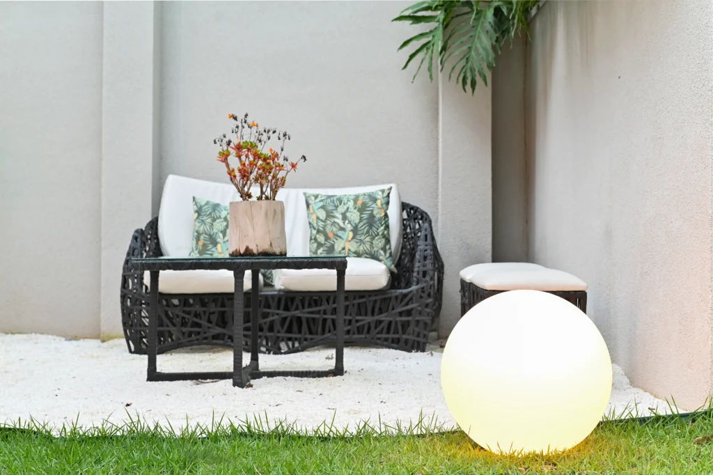 <img src="area-externa-varanda-movel-vime-preto-sofa-branco-luminaria-esfera.webp" alt="Área externa de varanda com mobília de vime preto, sofá branco com almofadas decorativas e luminária de esfera em ambiente ao ar livre com plantas e paredes claras.">