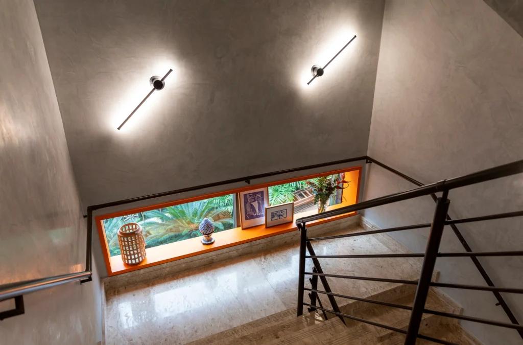 <img src="escada-interior-moderna-com-janela-decorada.webp" alt="Imagem do interior de uma escada com iluminação moderna, parede de cimento e uma janela panorâmica decorada com plantas e quadros, vista de cima.">