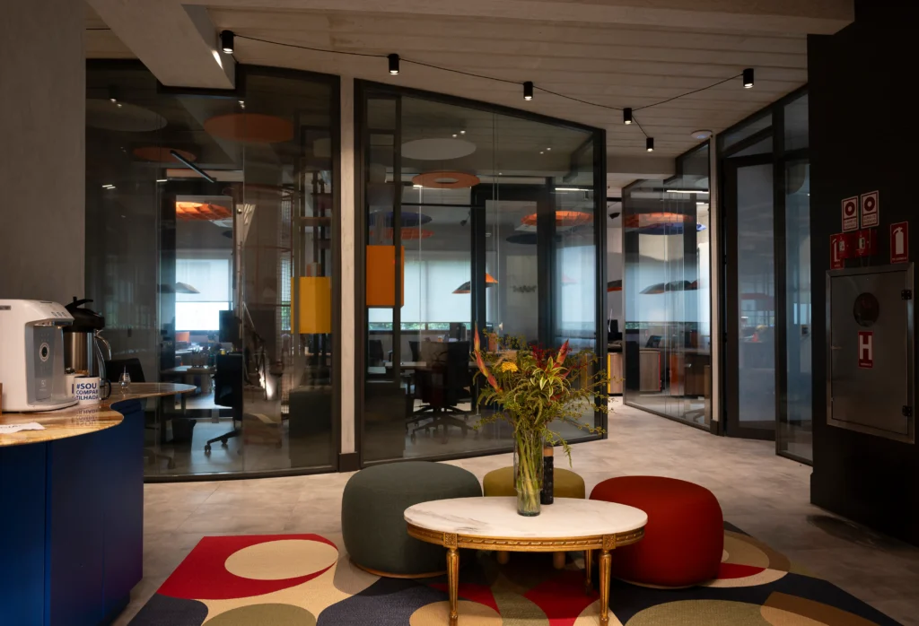 <img src="escritorio-moderno-area-convivencia.webp" alt="Imagem de um espaço de escritório moderno com área de convivência que inclui uma mesa decorada com vaso de flores, poltronas coloridas, e vidro divisas de ambientes de trabalho.">