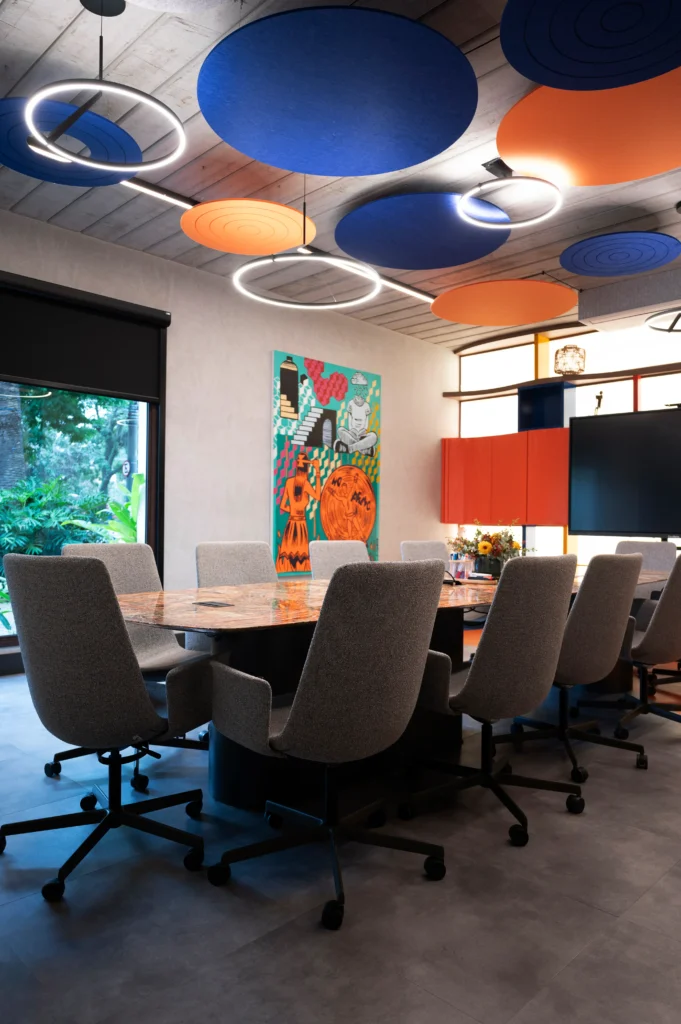 <img src="sala-de-reuniao-moderna-com-decoracao-colorida.webp" alt="Sala de reunião moderna com mesa de madeira e cadeiras confortáveis, decoração colorida com arte vibrante na parede, iluminação contemporânea no teto azul e laranja, e grande janela com vista para plantas verdes.">