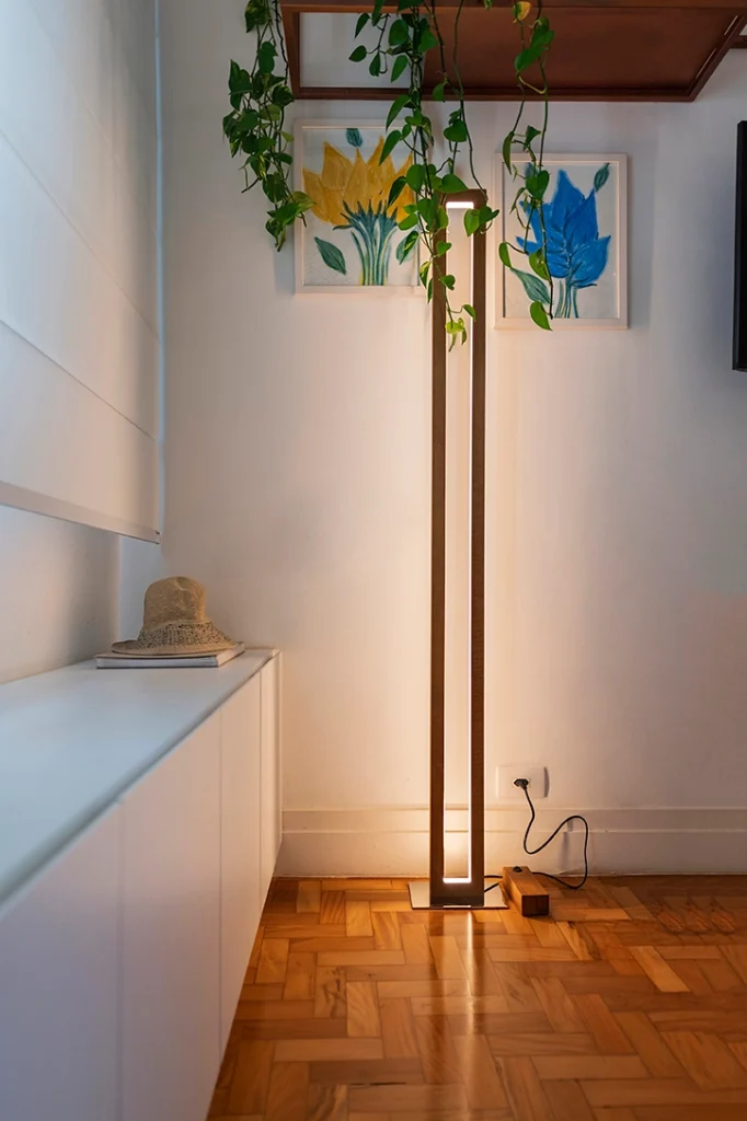 <img src="lampara-de-chao-moderna-decorativa-sala-flores-quadrinhos-plantas.webp" alt="Lâmpada moderna de chão acesa em sala com piso de madeira, decorada com quadros de flores coloridas e plantas penduradas no teto.">