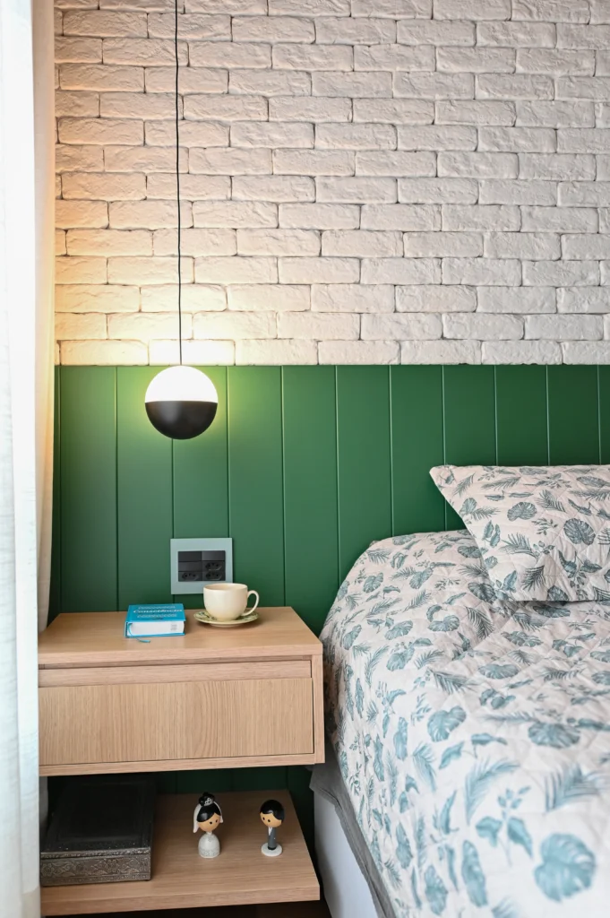 <img src="quarto-decoracao-moderna-parede-tijolos-verdes.webp" alt="Imagem de um quarto com parede de tijolos brancos na parte superior e painéis verdes na parte inferior, com iluminação moderna e acessórios de decoração, incluindo uma cama com roupa de cama floral, luminária pendente preta e branca, e uma mesa de cabeceira de madeira com livros, uma xícara e figuras decorativas.">