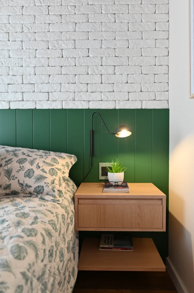 <img src="quarto-decoracao-parede-tijolos-brancos-cabeceira-verde.webp" alt="Quarto com parede de tijolos brancos, cabeceira verde escura, lâmpada de leitura preta ao lado da cama com roupa de cama com estampa de folhas verdes, e uma mesinha de cabeceira de madeira com uma planta, óculos e livros.">