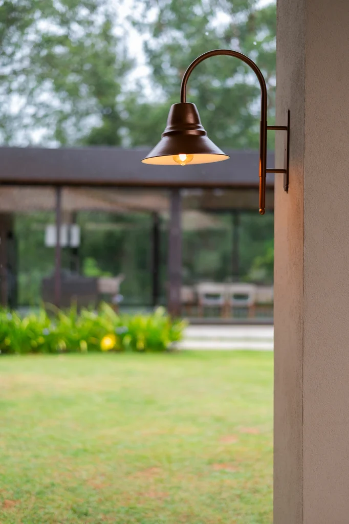 <img src="luminaria-parede-ferro-varanda-jardim.webp" alt="Luminária de parede de ferro lateral instalada na parede de uma varanda, com luz amarelada, em frente a um jardim verde e lateral de uma residência.">
