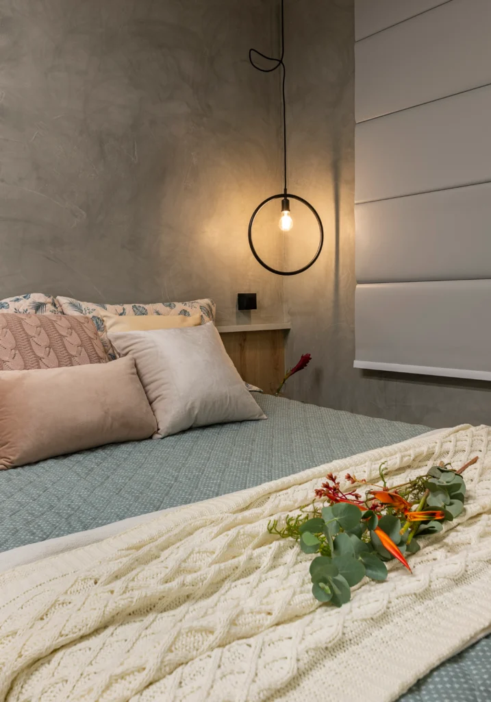 <img src="quarto-decorado-com-lampada-pendente-e-cama-de-casal-2023.webp" alt="Cama de casal decorada com almofadas bege, cobertor azul claro, em um quarto aconchegante com parede de cimento e iluminação com lâmpada pendente moderna.">