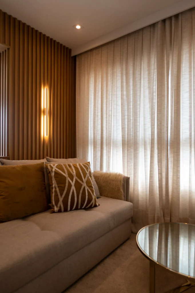 <img src="sala-de-estar-moderno-com-cortinas-e-sofa-bege-jpg.webp" alt="Imagem de uma sala de estar moderna com cortinas leves, sofá bege decorado com almofadas douradas e uma parede de madeira ressaltando uma iluminação de destaque.">