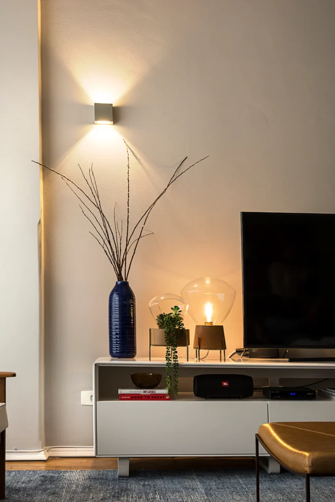 <img src="decoracao-de-sala-modernizada-com-vaso-decorativo-e-ambiente-confortavel.webp" alt="Decoração de sala moderna com luminária de parede, vaso com galhos secos, luminárias de vidro e TV, criando um ambiente aconchegante e elegante.">