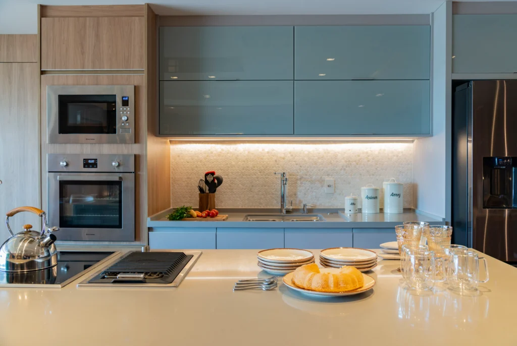 <img src="cozinha-modernaconfortavel-organizada.webp" alt="Cozinha moderna e organizada com armários azuis, eletrodomésticos integrados, bancada com pratos, copos e utensílios, iluminação revestida de mármore.">