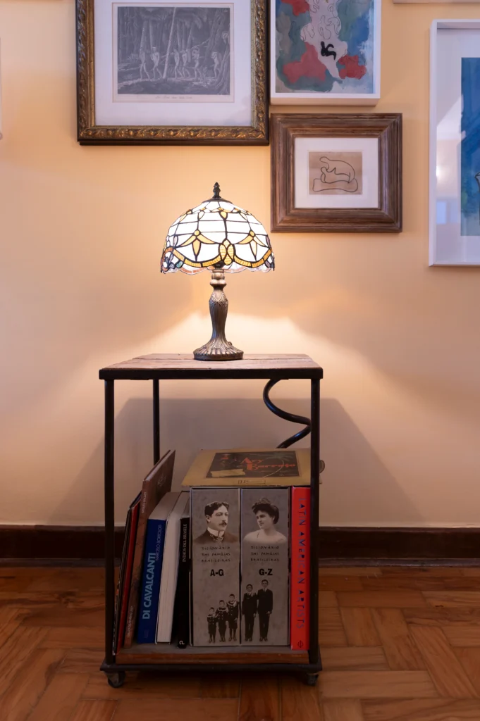 <img src="lampara-tiffany-ambiente-de-arte.webp" alt="Lâmpada de mesa de estilo Tiffany iluminando uma pequena mesa de madeira com prateleira, ladeada por quadros de arte moderna na parede amarela.">