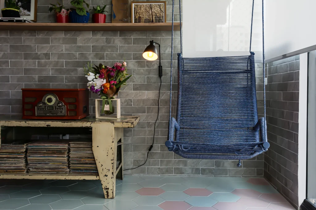 <img src="decoracao-varanda-poltrona-corda-azul.webp" alt="Decoração de varanda com poltrona de corda azul, mesa de madeira vintage com pilhas de vinis, luminária de parede e plantas decorativas em ambiente moderno">