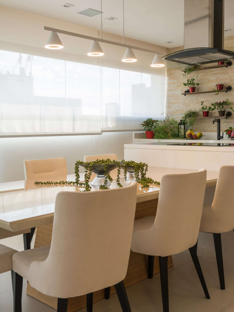 Sala de jantar moderna com mesa retangular de madeira clara e cadeiras estofadas em tom bege. Sobre a mesa, um elegante arranjo de plantas verdes. O ambiente é iluminado por três pendentes contemporâneos, com lâmpadas de luz suave, destacando o estilo minimalista do espaço. Ao fundo, cozinha integrada com balcão branco e prateleiras com vasos de plantas decorativas