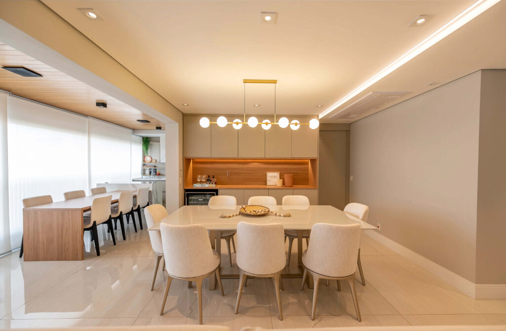 Sala de jantar moderna com mesa retangular clara e cadeiras estofadas em tom bege. Acima da mesa, uma luminária contemporânea com esferas brancas e estrutura metálica dourada, iluminando suavemente o ambiente. Ao fundo, uma área de bar com armários em tons neutros e detalhes em madeira, criando um contraste elegante. O espaço é complementado por iluminação embutida no teto e piso de porcelanato claro, proporcionando um ambiente sofisticado e acolhedor