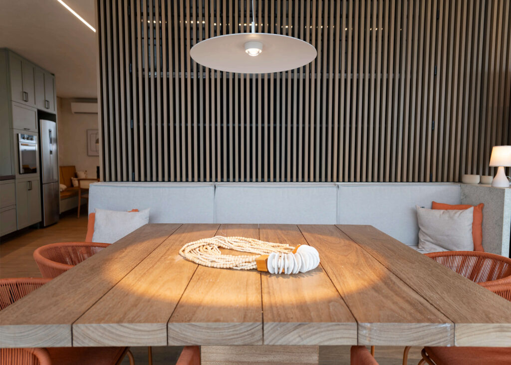 Sala de jantar com mesa de madeira de design rústico, iluminada por um pendente moderno de formato circular e cor branca, que destaca o centro da mesa. As cadeiras de fibra laranja complementam a decoração contemporânea. Ao fundo, uma parede com ripas de madeira cria um ambiente sofisticado, contrastando com o sofá claro e almofadas coloridas