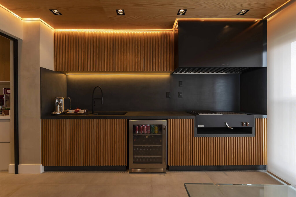 Área gourmet moderna com armários de madeira, refrigerador e iluminação embutida em LED, destacando a importância da iluminação na criação de um ambiente sofisticado e funcional