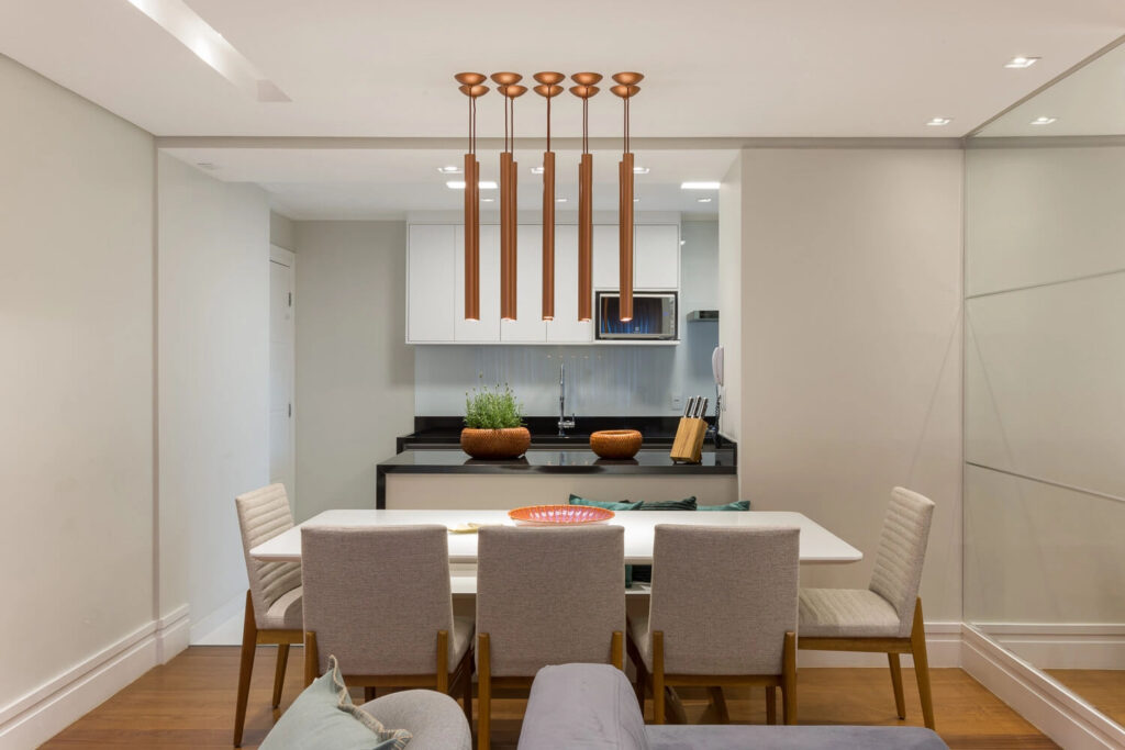 Sala de jantar moderna com mesa retangular branca e quatro cadeiras claras. Sobre a mesa, um pendente de design contemporâneo em cobre, composto por lâmpadas lineares pendentes, proporcionando uma iluminação suave e elegante para ocasiões especiais como festas de fim de ano. Ao fundo, uma cozinha integrada com armários brancos e balcão de granito preto