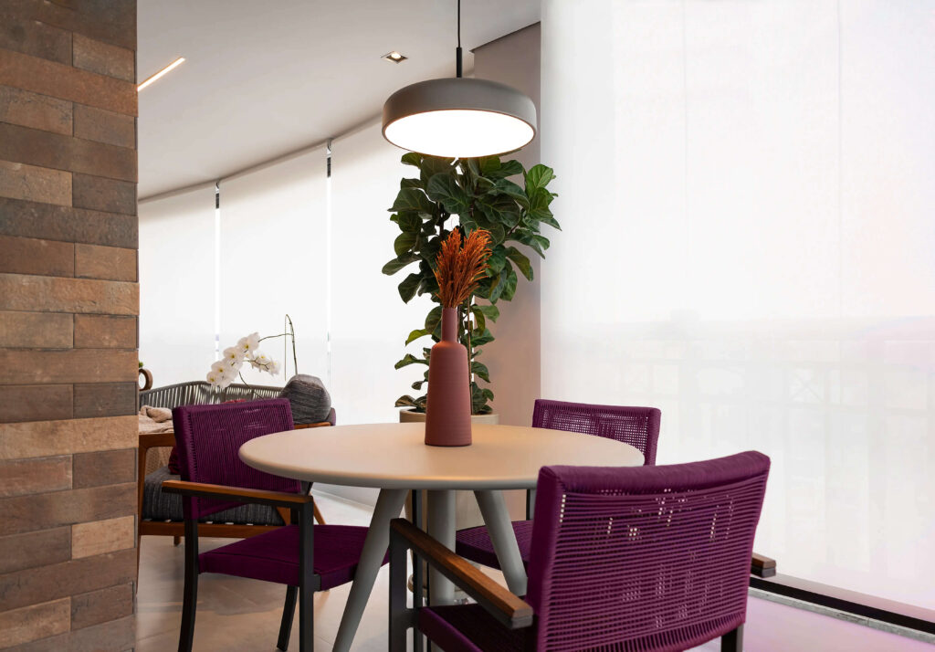 Sala de jantar com mesa redonda clara e cadeiras roxas de design moderno. No centro da mesa, um vaso simples com flores secas. Acima, uma luminária pendente de formato circular e cor metálica ilumina o ambiente de maneira suave. Ao fundo, uma planta grande adiciona um toque de verde, complementando a decoração contemporânea e acolhedora do espaço