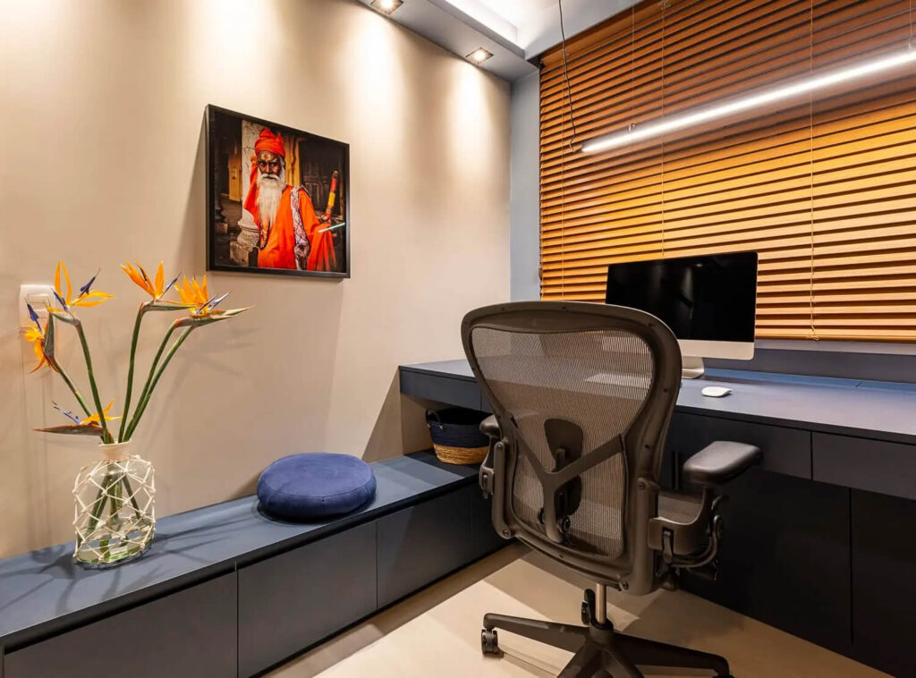 Um cantinho moderno e tranquilo para leitura ou trabalho, com uma cadeira ergonômica confortável e uma mesa minimalista. A iluminação é composta por spots direcionáveis no teto, proporcionando luz focada e suave, além de uma luminária sobre a mesa que ilumina de forma suave e aconchegante. Ao fundo, uma janela com persiana de madeira e um vaso com flores tropicais, criando um ambiente acolhedor e inspirador.